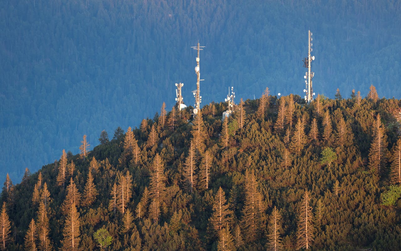 A serene mountain peak with radio antennas surrounded by dense forest during sunset.